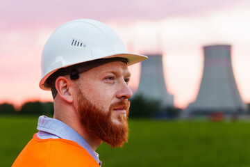 Portrait of a nuclear power plant engineer. A man with a beard in a protective helmet and an orange vest stands against the background of a nuclear power plant