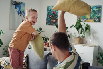 Happy little son, playing pillow fight together with his dad on sofa during leisure time at home
