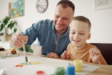 Happy father painting a picture with paintbrush together with his little son at table