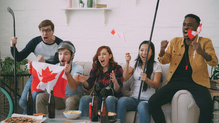 Group of young friends sports fans with Canadian national flags watching sport championship on TV together cheering up favourite team at home indoors - 515574877