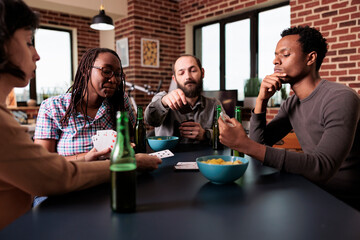 Multicultural group of friends sitting at table in living room while playing guessing card game together. Multiethnic young adult people at home enjoying fun leisure activity.