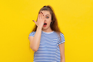 Portrait of surprised brunette girl, cover half of face and open mouth, looking at camera with one eye, checking vision, standing in white-blue striped t shirt over yellow background