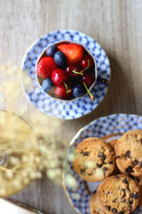Plate of chocolate chip cookies, cup filled with strawberries, blueberries and cherries, open book and vase with gypsophila flowers on the table. Flat lay.