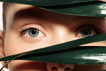 Closeup young beautiful woman's face with healthy skin. Blue eyes looking through palm leaves. Model with blue eye and without makeup. Skin care concept.