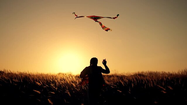 Happy child. Girl with flying kite in park. Child runs at sunset on grass in park. Silhouette of girl with flying kite. Child launches flying kite into sky in the wind. Silhouette of child in meadow