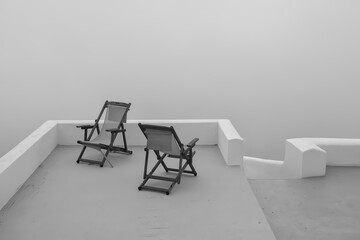 View of two empty sunbeds in front of the aegean sea that is covered by fog in Santorini Greece in black and white 