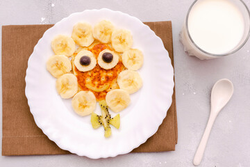 Funny chamomile flower face shape snack from pancake,bananas,kiwi.Cute kids childrens baby's sweet dessert,healthy breakfast,lunch,food art with milk on plate,gray concrete background,top view