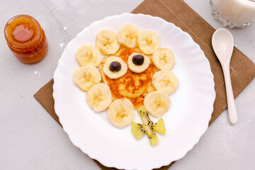 Funny chamomile flower face shape snack from pancake,bananas,kiwi.Cute kids childrens baby's sweet dessert,healthy breakfast,lunch,food art with milk,honey on plate,gray concrete background,top view