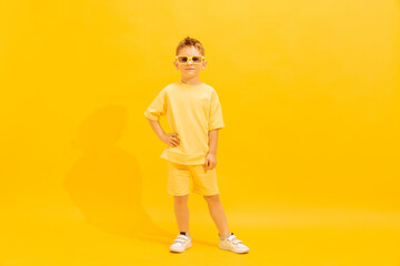 Fototapeta na wymiar Portrait of cheerful little boy, child in bright summer outfit and sunglasses posing isolated over yellow background