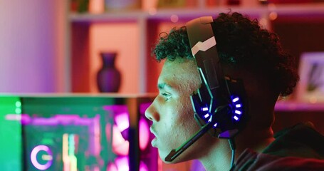 Guy playing video games on a computer with neon lights at night. Closeup of a focused esports gamer wearing a headset to chat to friends while streaming and competing in a multiplayer online session