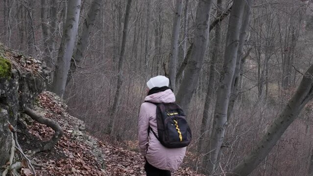 Woman with white cup and gree jaket, wearing backpack walks downhill through a forest. Lost in the woods concept.