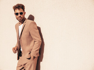 Handsome confident stylish hipster lambersexual model.Sexy modern man dressed in elegant beige suit. Fashion male posing in the street background near white wall at sunset. In sunglasses