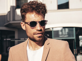 Handsome confident stylish hipster lambersexual model.Sexy modern man dressed in elegant beige suit. Fashion male posing in the street background in Europe city at sunset. In sunglasses