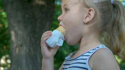 Fototapeta premium Cute little girl eats ice cream outside. Close-up portrait of blonde girl sitting on park bench and eating icecream.
