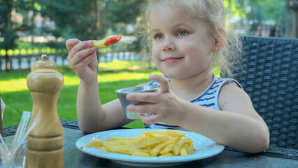 Little girl eat french fries. Close-up of blonde girl takes potato chips with her hands and tries them sitting in street cafe on the park.