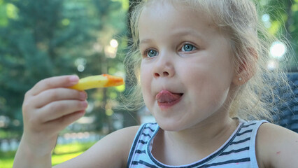 Little girl eat french fries. Close-up of blonde girl takes potato chips with her hands and tries them sitting in street cafe on the park.