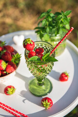 Fresh homemade lemonade with strawberries and peppermint in green glass with straw on white round table outdoors in sunny summer day. Healthy lifestyle