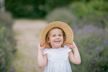 Portrait of a cute little curly blue-eyed girl 3 years old in a wicker hat and white sundress near...