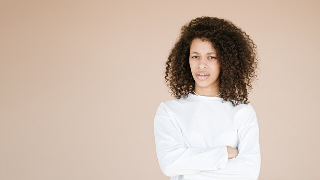 Displeased curly woman looks angrily at camera, smirks and looks from eyebrows, offended by boyfriend, focused at camera, standing upbeat against brown background       