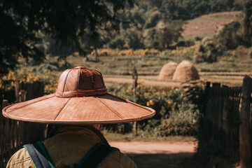 Person with a large bamboo hat walking in the countryside with fields and haystacks in the blurry background.