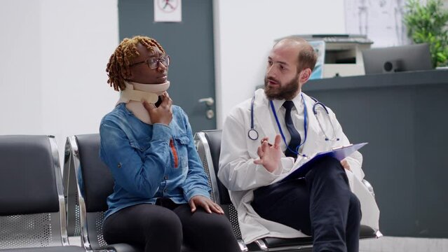 Female patient with cervical neck foam talking to doctor in waiting area at healthcare facility. Hurt woman in pain with collar brace trying to cure physical injury, help from medic. Handheld shot.