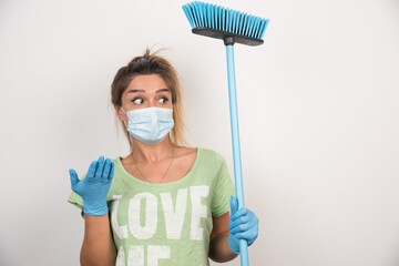 Young housewife with facemask and broom pointing sideways with her hand on white background
