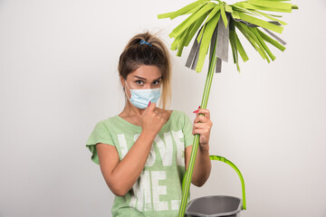 Young housewife with facemask holding mop and looking front on white background