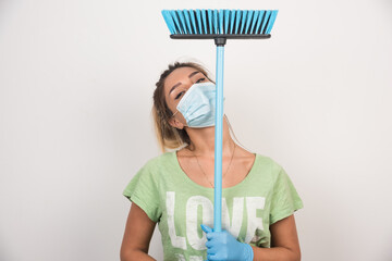 Young housewife with facemask holding broom while looking front on white background