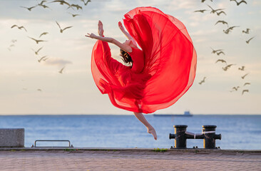 Jumping ballerina in a red flying skirt and leotard on ocean embankment or sea beach surrounded by...
