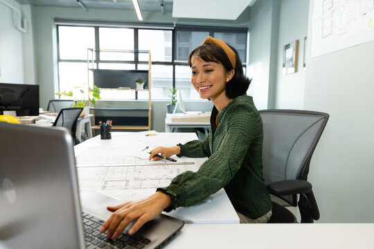 Smiling asian young female architect with blueprint using laptop at table in office