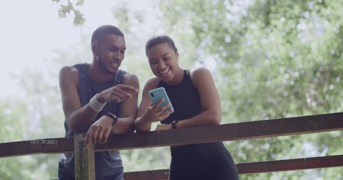 Two Joggers Using Their Phone In A Park And Looking At A Fitness App During A Break. Happy Athletic Couple Discussing Exercise Goals Or Searching For Hiking Trails In Remote Location With Online GPS