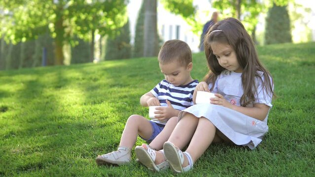 Two children, a boy and a girl, are sitting on the lawn in the park on a bright sunny summer day and eating ice cream with a spoon. Happy childhood in the fresh air. Children smile and enjoy the treat