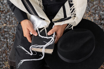 Close-up of a Jew holding a siddur on which rests the tzitzit of his tallit and next to it a hat. - obrazy, fototapety, plakaty