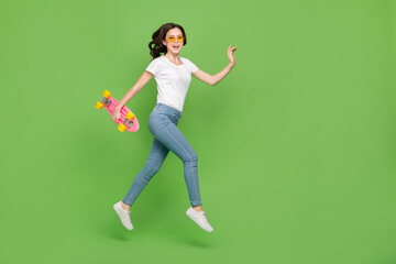 Fototapeta na wymiar Photo of excited funny woman dressed white t-shirt eyewear holding skate board jumping empty space isolated green color background