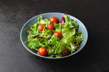 Green salad leaves with cherry tomatoes in bowl on black table.