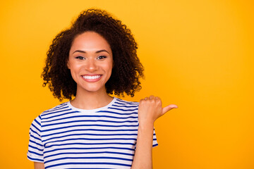 Photo of pretty cheerful girl dressed striped t-shirt pointing thumb empty space isolated yellow...
