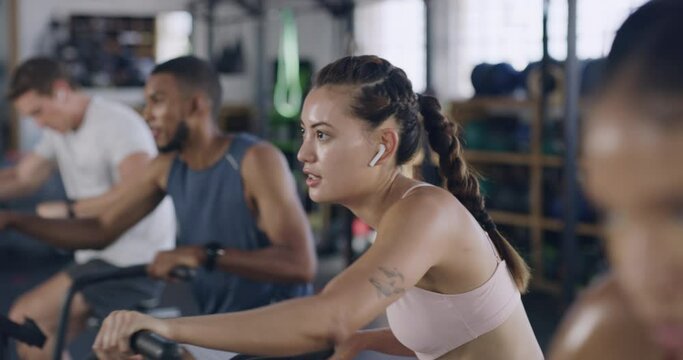 Focused and sporty woman during a spinning class at the gym. Fit people using air bikes for cardio exercise in a fitness facility. Group of sweaty men and women listening to music while exercising