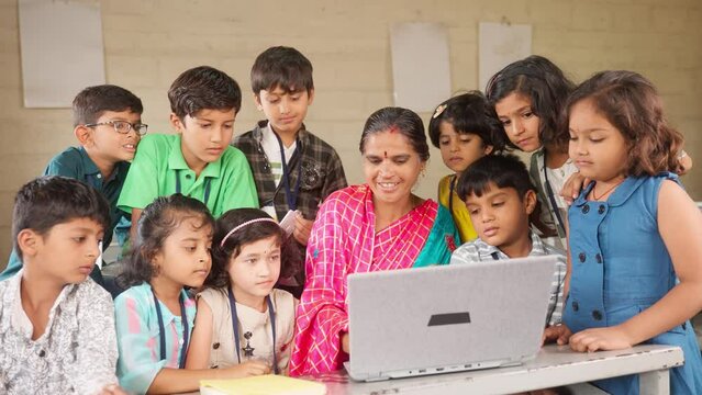 Happy teacher teaching from laptop to elementary students around at classroom - concept of development, technology and learning