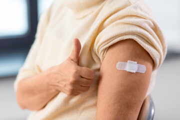 medicine, health and vaccination concept - close up of vaccinated senior woman in mask with medical patch on arm showing thumbs up at hospital