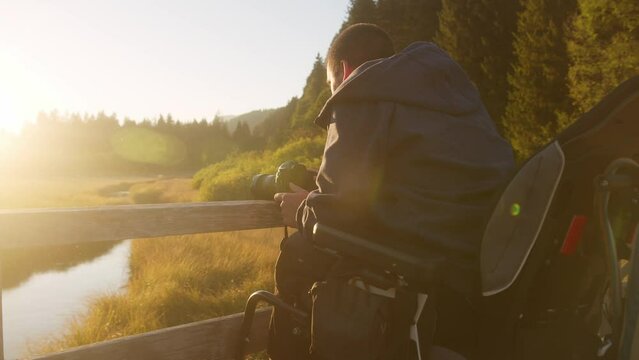 Young Caucasian man on wheelchair on a wooden bridge, using a camera to take a photo of beautiful mountain nature, handheld shot.