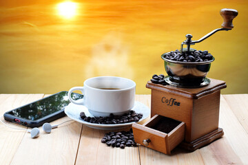 Coffee beans with a coffee grinder with a hot white coffee cup and mobile phone placed on a wooden floor with a yellow sky in the evening and the sun as the background. Close up. - Powered by Adobe