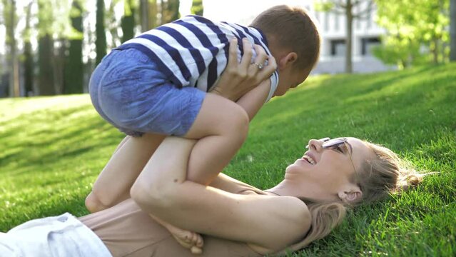 Mom lying on the grass in the park smiling lifts her son up on a bright sunny day. Family video. The girl lifts the child above her lying on the grass in the park at sunset. Happy childhood