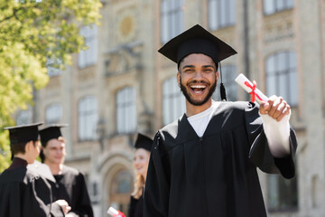 Happy african american bachelor in gown holding diploma outdoors.