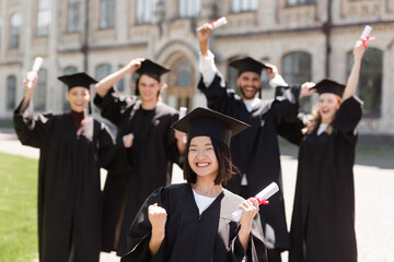 Excited asian bachelor holding diploma near blurred friends in park.