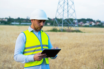 a male engineer in a helmet and goggles uses a smartphone for field work near a telecommunications tower that controls cellular electrical installations.