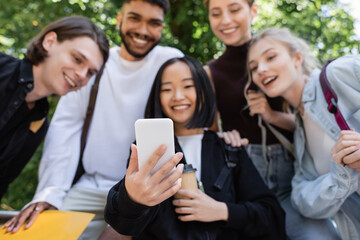 Blurred asian student holding smartphone near interracial friends in park.