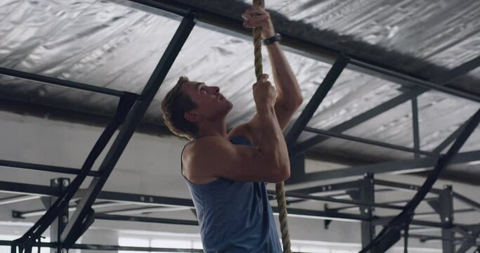 Strong, active, athletic trainer using upper body strength and strong arms to pull himself up in a HIIT fitness workout. Focused on muscle health. Fit man climbing rope for training exercise in a gym