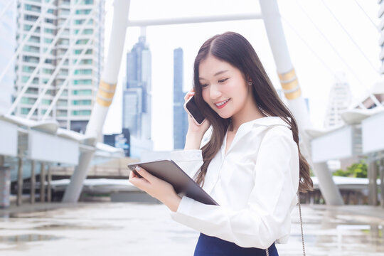 Professional business Asian female is walking to office or work place while she holds tablet in her hand and talks to customer on the phone with smartphone at outdoors among business buildings