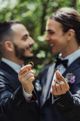 happy and blurred gay men holding wedding rings in hands and looking at each other.