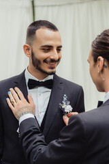 tattooed gay man adjusting boutonniere on suit of happy bearded groom.